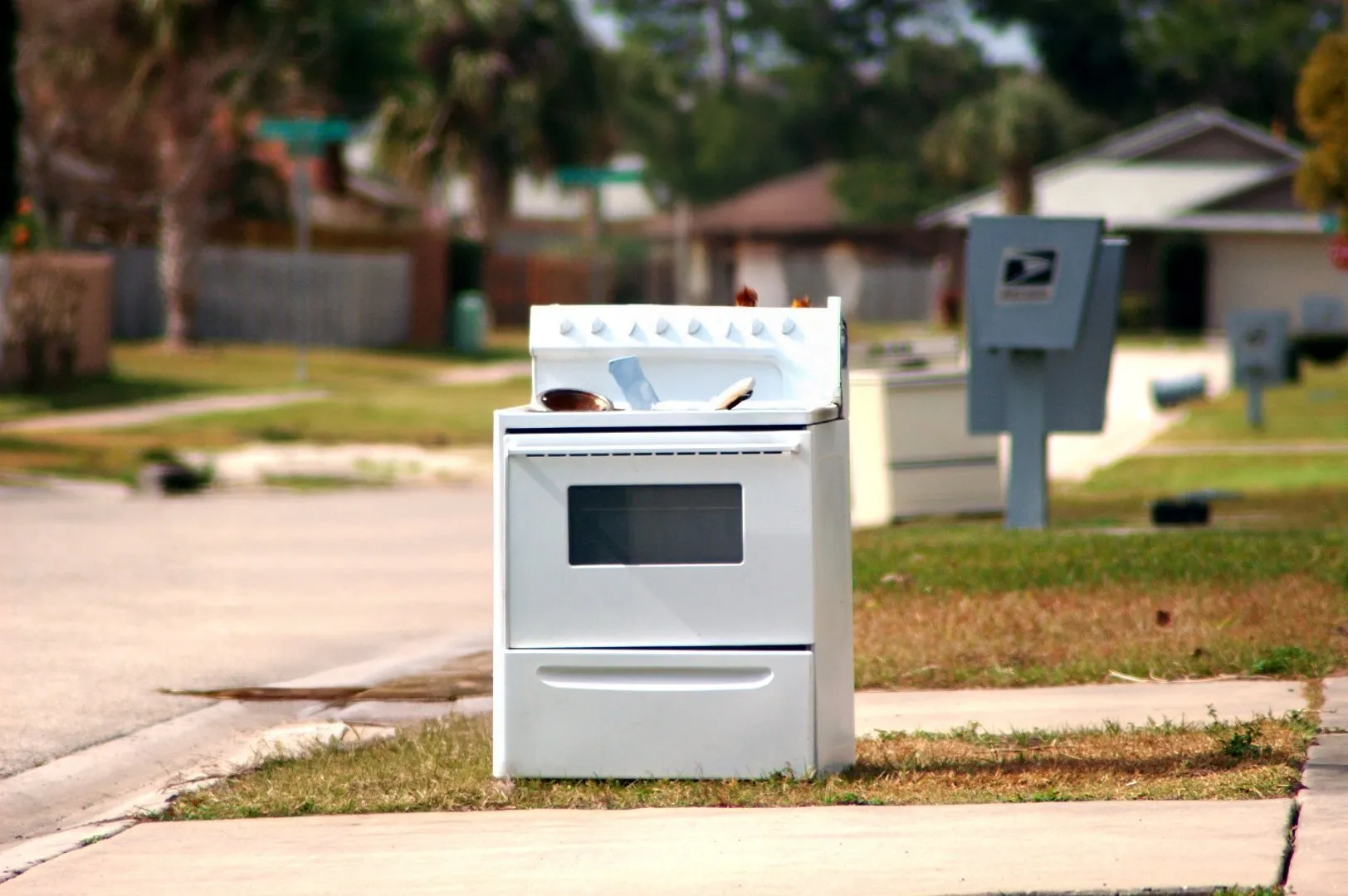White electric stove with four burners and an oven placed on a curbside next to a sidewalk in a residential neighborhood, positioned for disposal or bulk pickup. Mailboxes and houses are visible in the background under sunny weather.