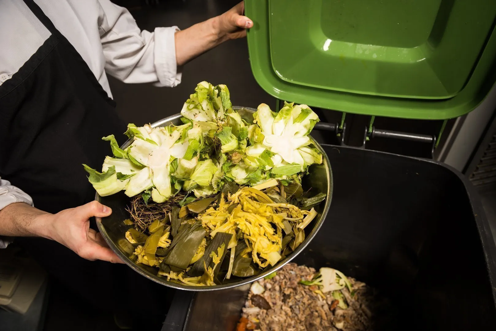Chef in a white coat holds a bowl of vegetable scraps over a green-lidded compost bin, with additional food waste visible inside the container.