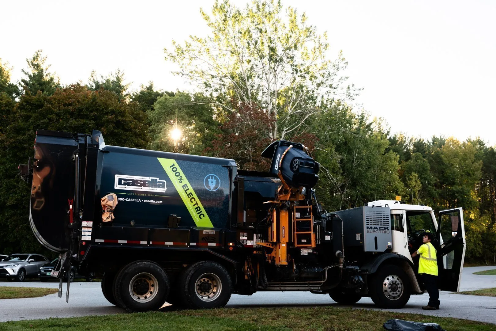 Casella's electric waste collection truck servicing a waste container near a grassy area.
