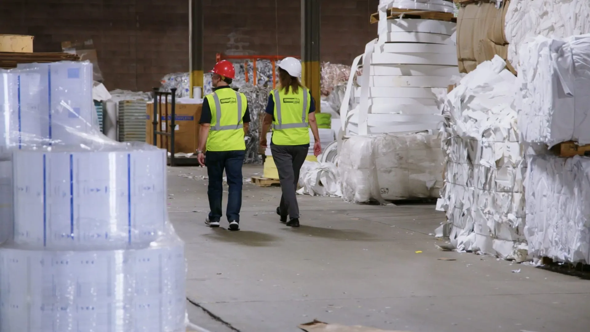 Two workers wearing bright yellow high-visibility safety vests and hard hats walk through an industrial warehouse filled with large stacks of baled white paper and rolls of plastic film. The concrete floor is clear, and the background includes metal shelving and bundled materials under bright overhead lighting.