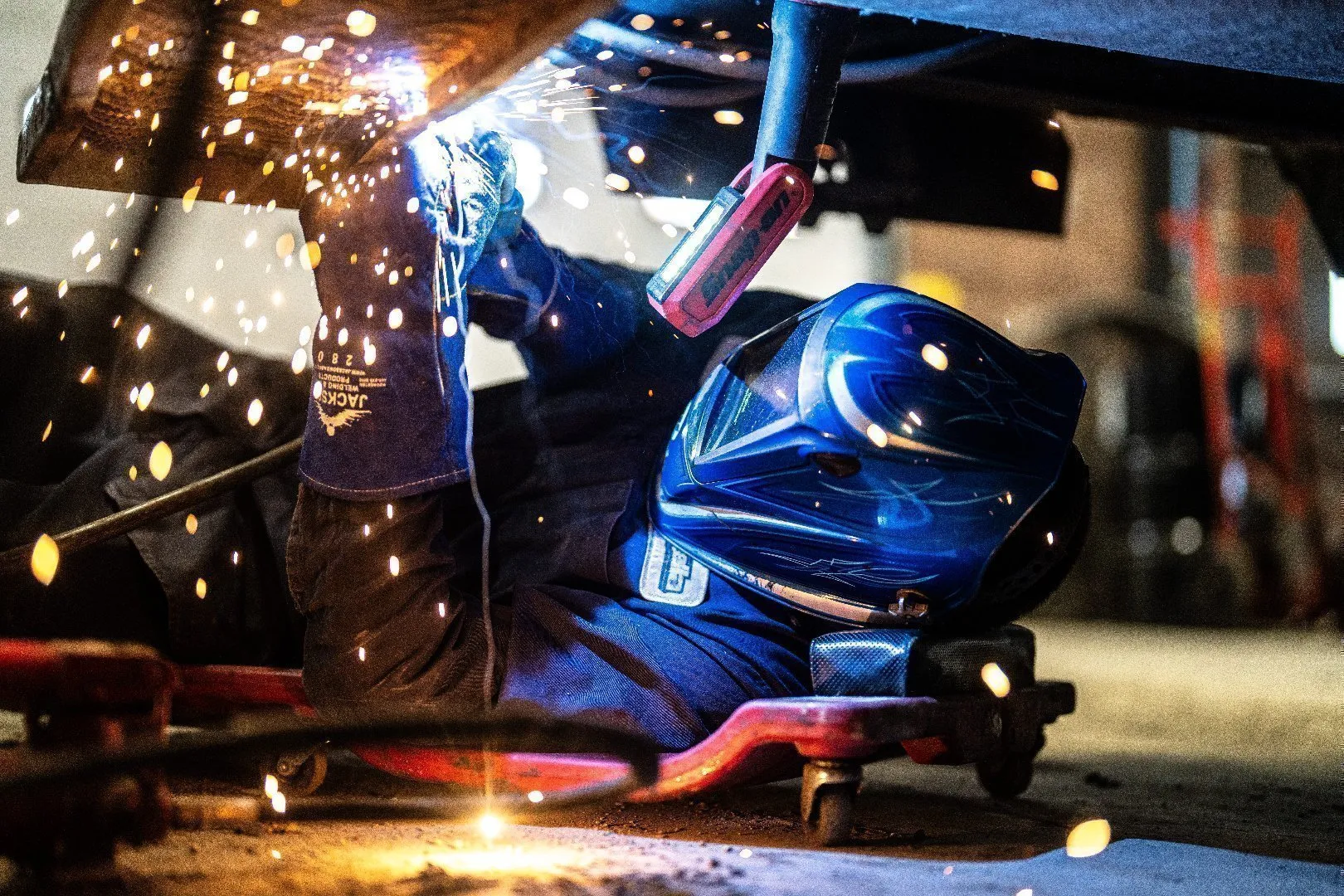 Technician wearing a blue welding helmet and protective gear uses a welding torch to repair metal underneath a large piece of equipment, with bright sparks flying across the workspace.