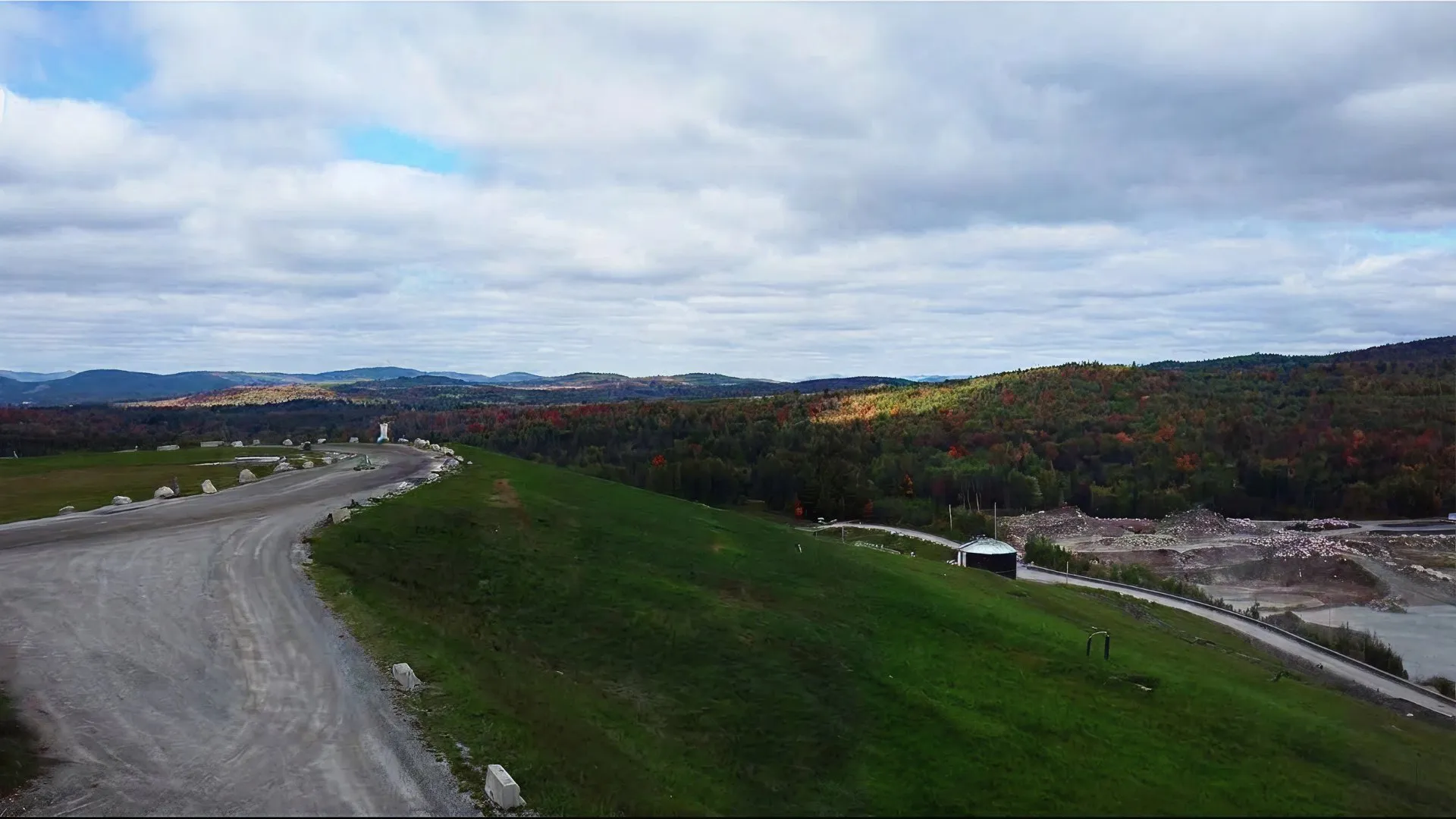 Landfill site with a dirt road overlooking forested hills under a cloudy sky.