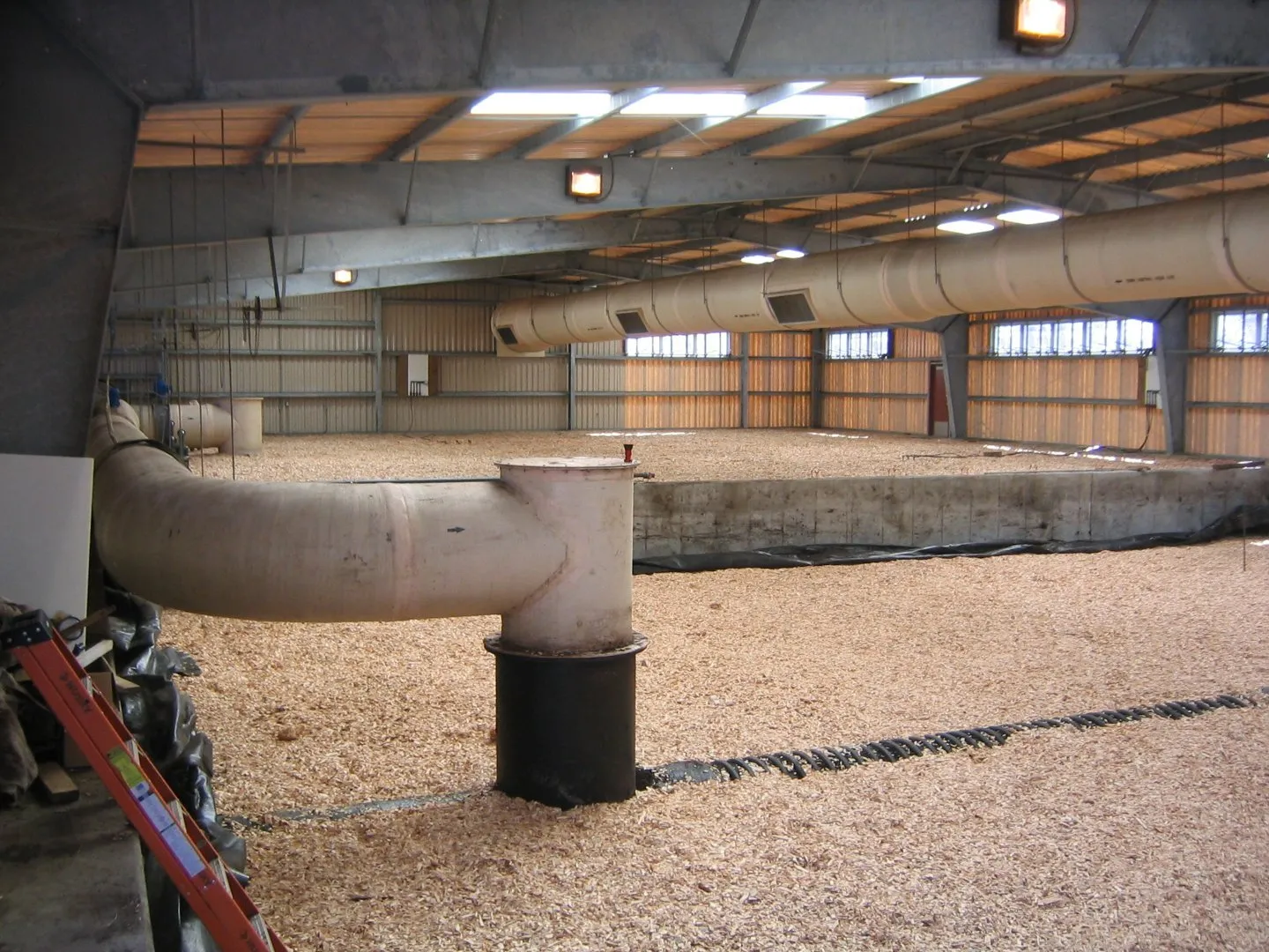 Indoor composting or processing facility with a large bed of wood chips, ventilation ducts overhead, and piping equipment in the foreground.