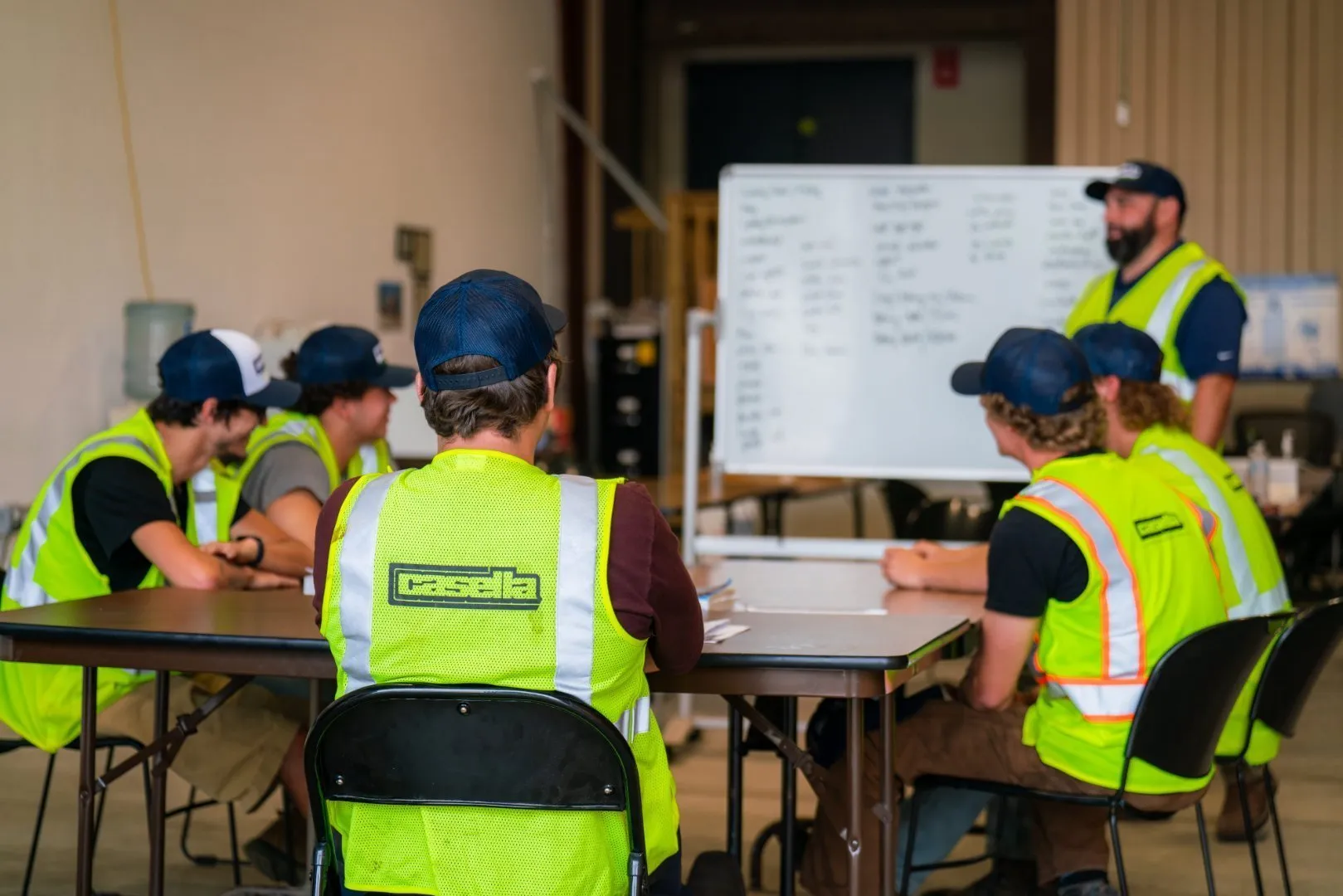 Group of students wearing bright yellow high-visibility safety vests and navy blue caps sit around tables in a training room, listening to an instructor standing near a large whiteboard filled with handwritten notes.