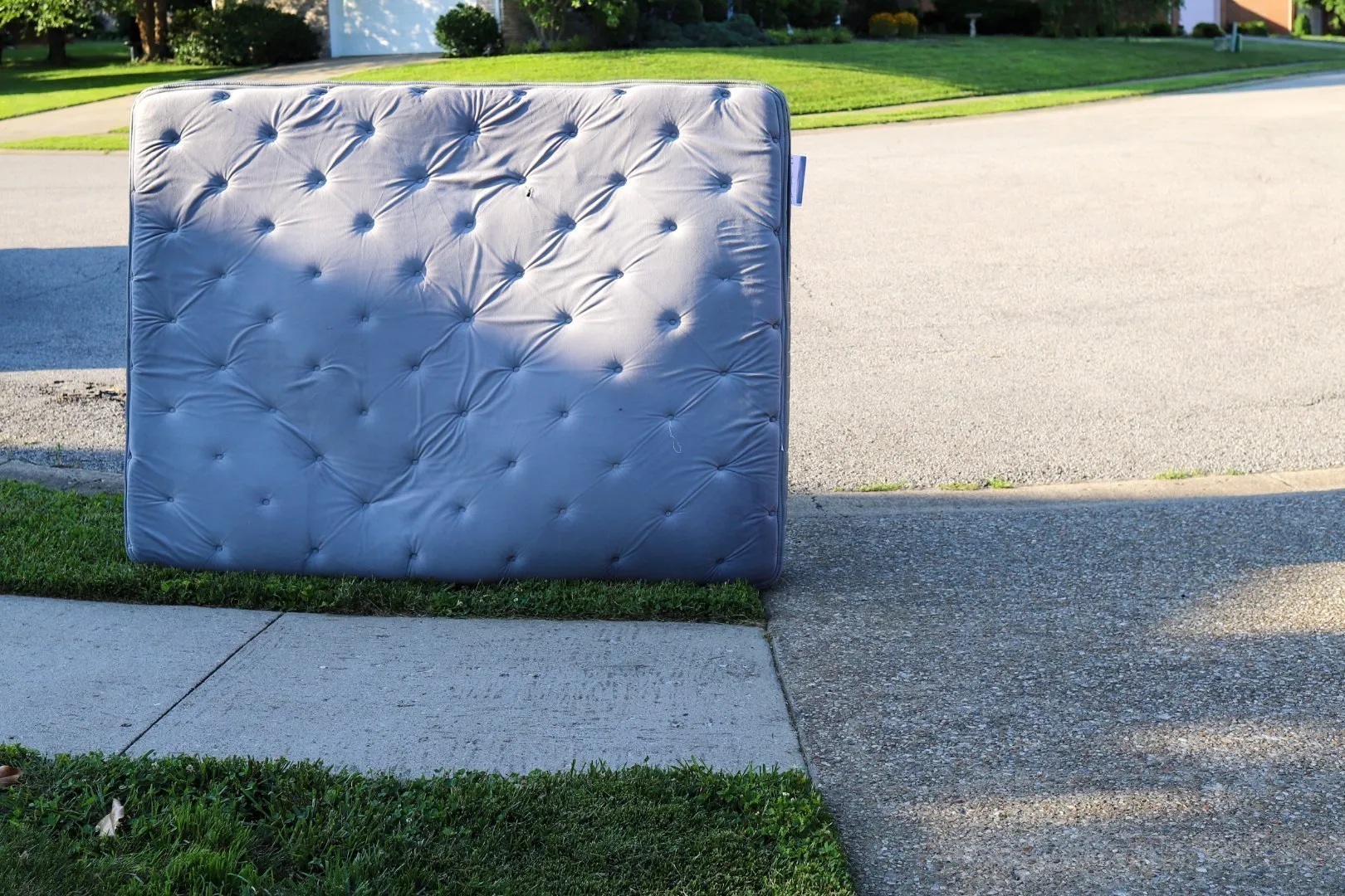 Large gray mattress with tufted design placed upright on a curbside next to a sidewalk and driveway in a residential neighborhood with green lawns and trees in the background.