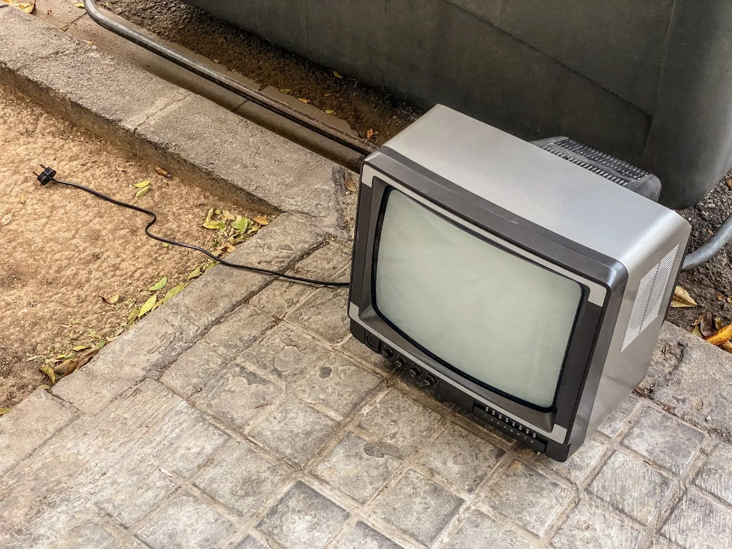 Old television with a gray casing and attached power cord placed on a tiled sidewalk next to a curb, positioned for disposal or recycling.