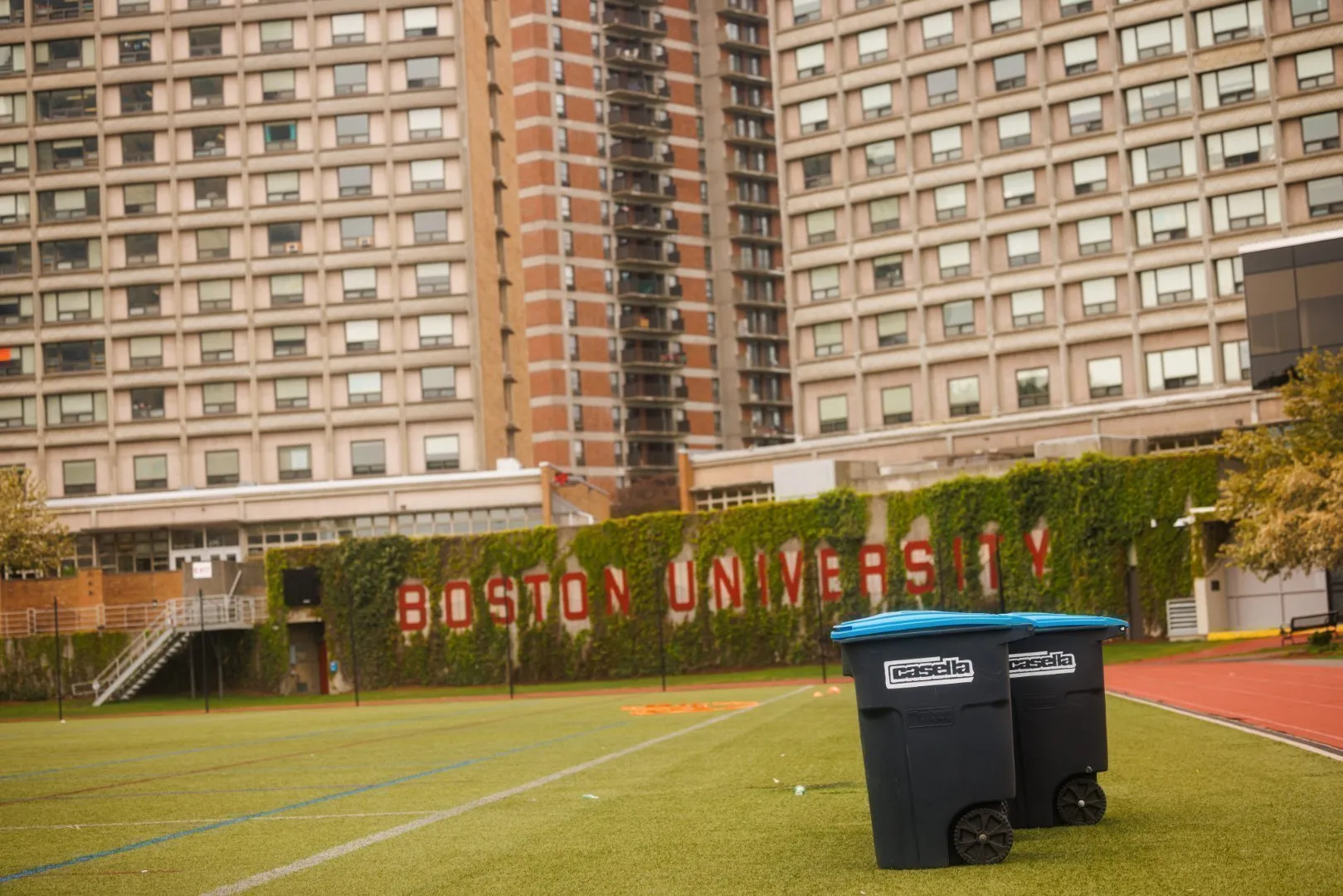 Two Casella recycling carts with blue lids placed on a green athletic field in front of a large brick building covered in ivy with the words “Boston University” displayed in bold red letters. Tall residential-style windows and multiple stories of the building are visible in the background under an overcast sky.