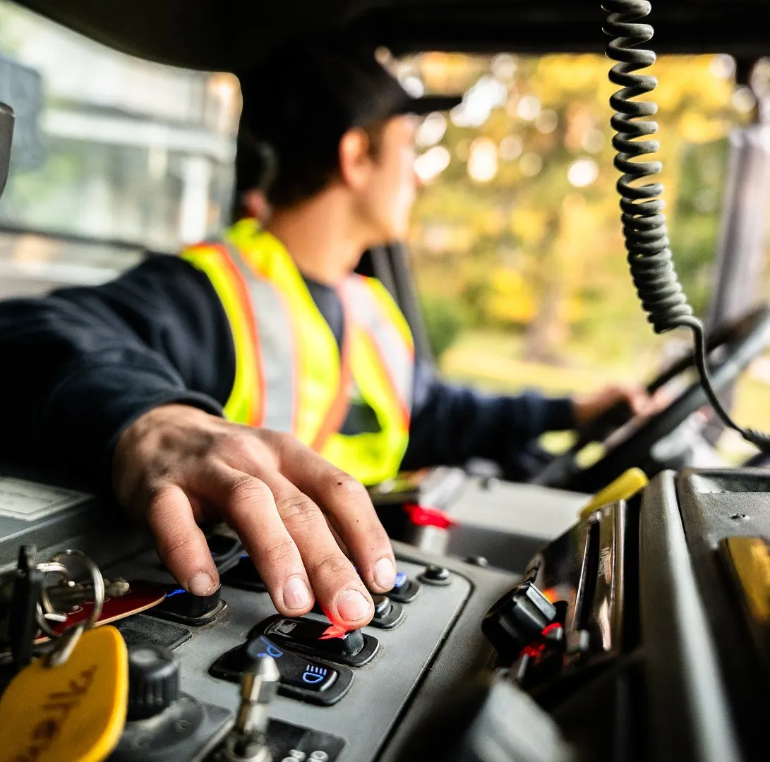 Truck driver operating dashboard controls inside a vehicle cab, wearing a reflective safety vest.