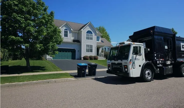 Casella garbage truck parked on a residential street in front of a two-story house with a green garage door and two blue curbside bins. 