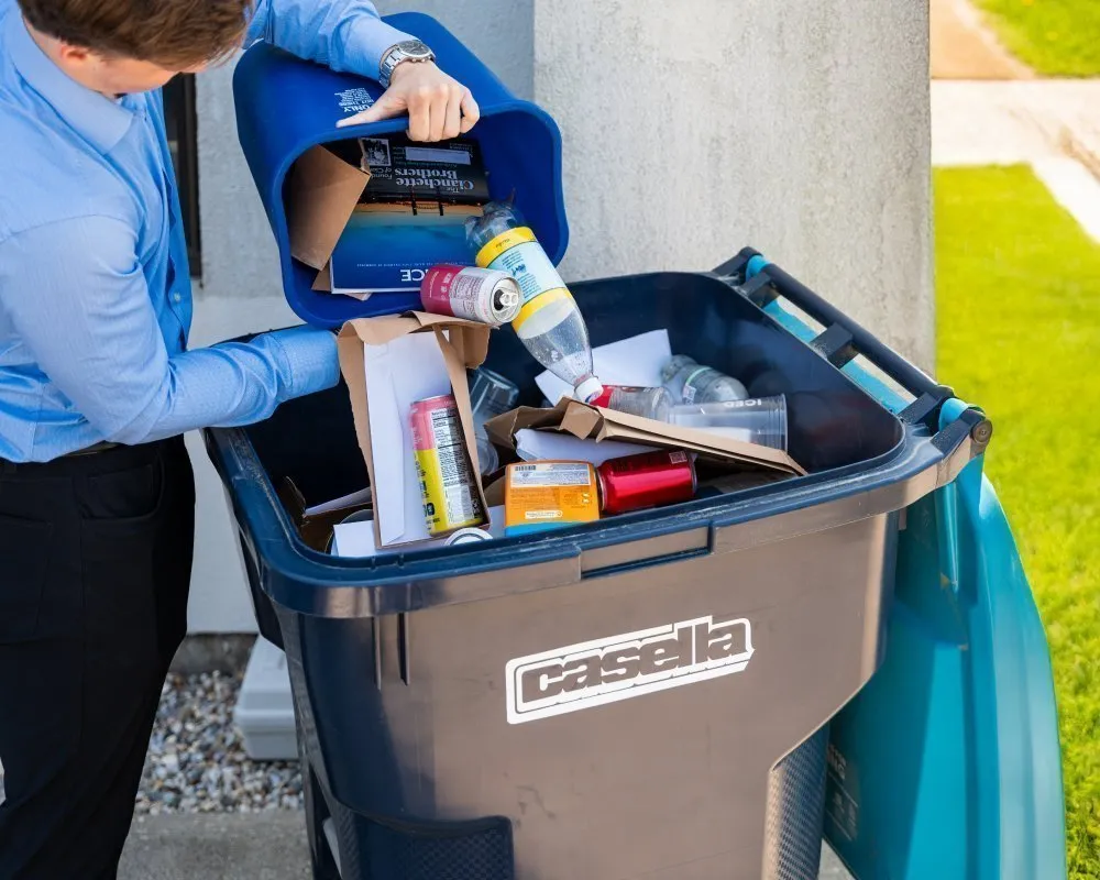 Person wearing a light blue long-sleeve shirt empties a small blue recycling bin into a large Casella-branded curbside recycling cart outdoors. The cart contains mixed recyclables including cardboard, paperboard boxes, aluminum cans, and plastic bottles. The setting includes a concrete wall, green grass, and a paved walkway under bright daylight. 