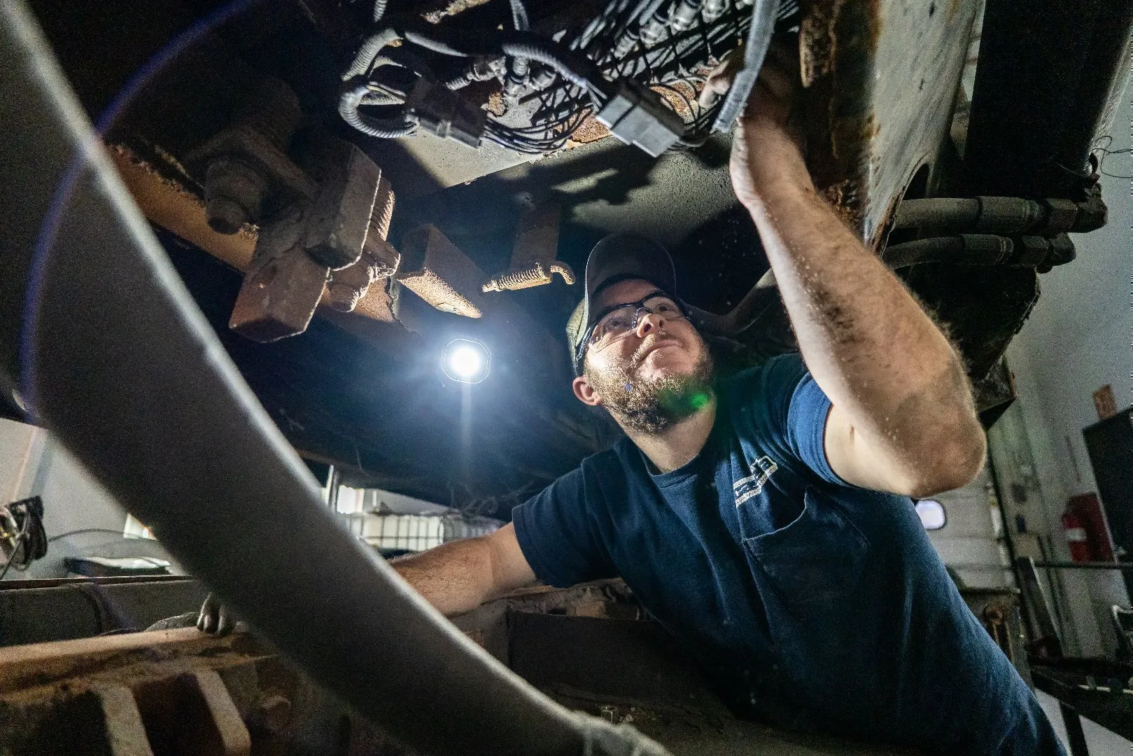 Diesel mechanic wearing a dark shirt and cap works underneath heavy equipment inside a workshop, inspecting wiring and components with a bright work light illuminating the area.
