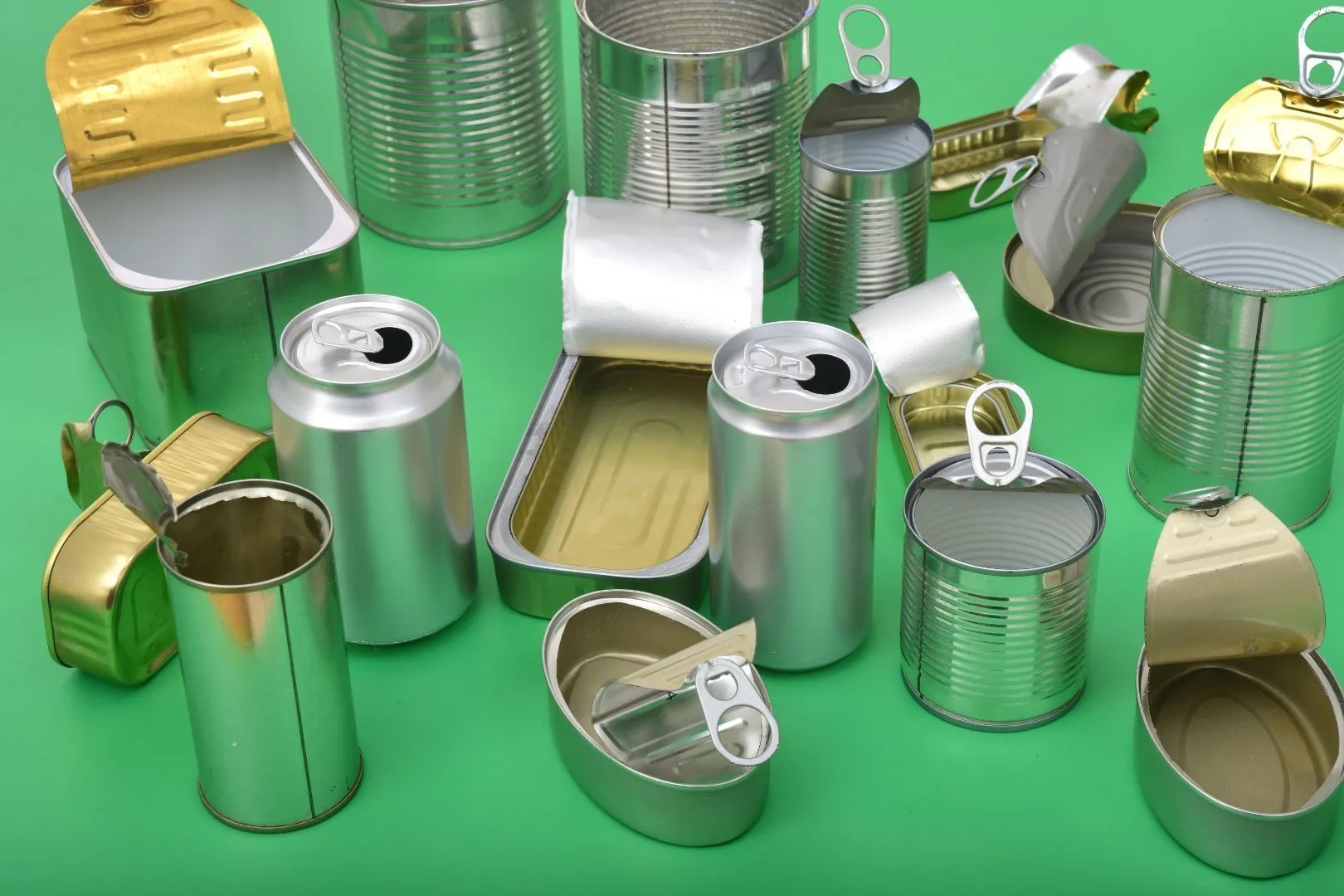 Collection of empty metal cans and aluminum containers of various shapes and sizes arranged on a bright green surface. Items include cylindrical food cans, beverage cans with pull tabs, and rectangular tins with gold and silver lids, some opened and others partially crushed. 
