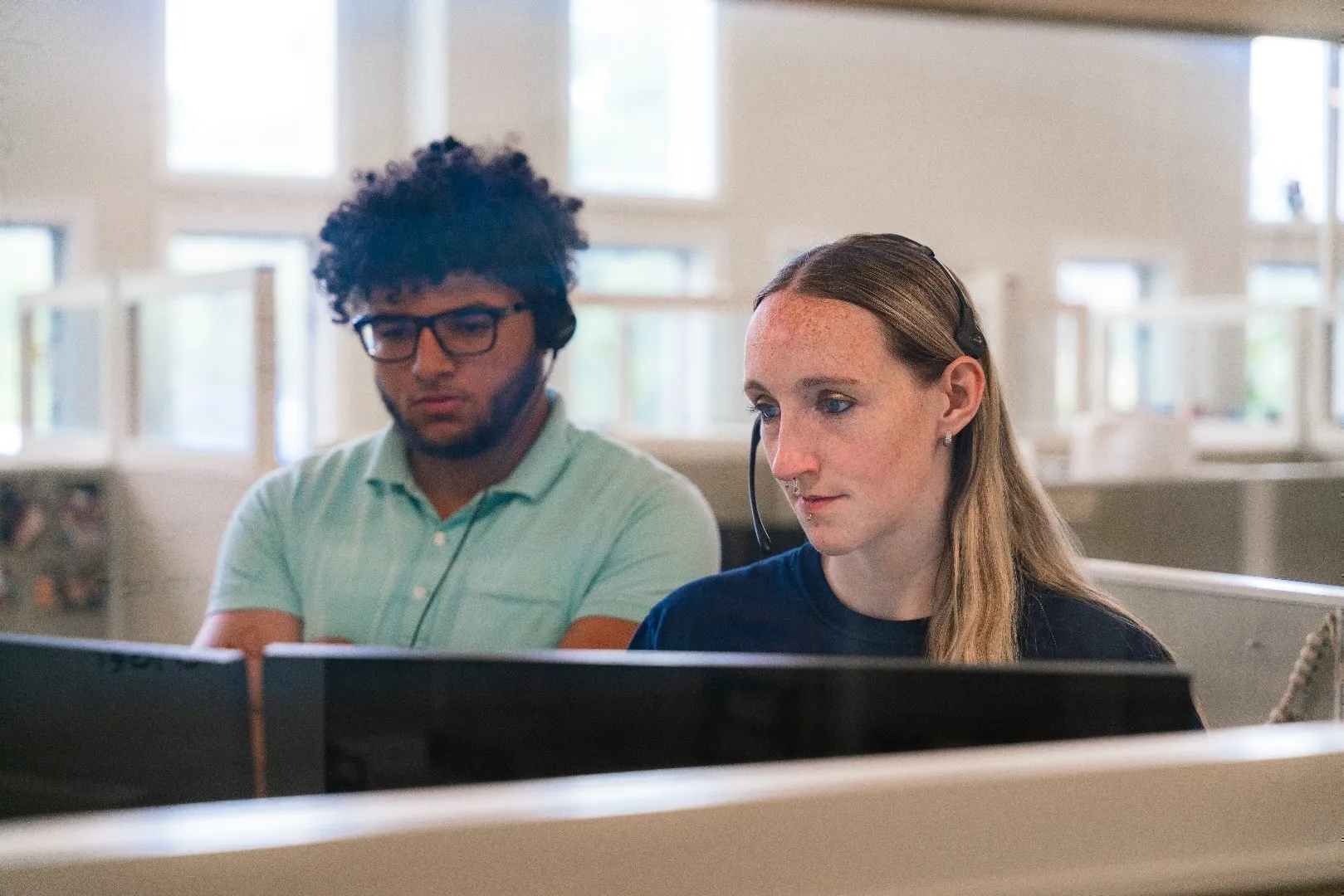 Two customer care representatives with headsets, in a modern office environment, working together on a computer monitor.
