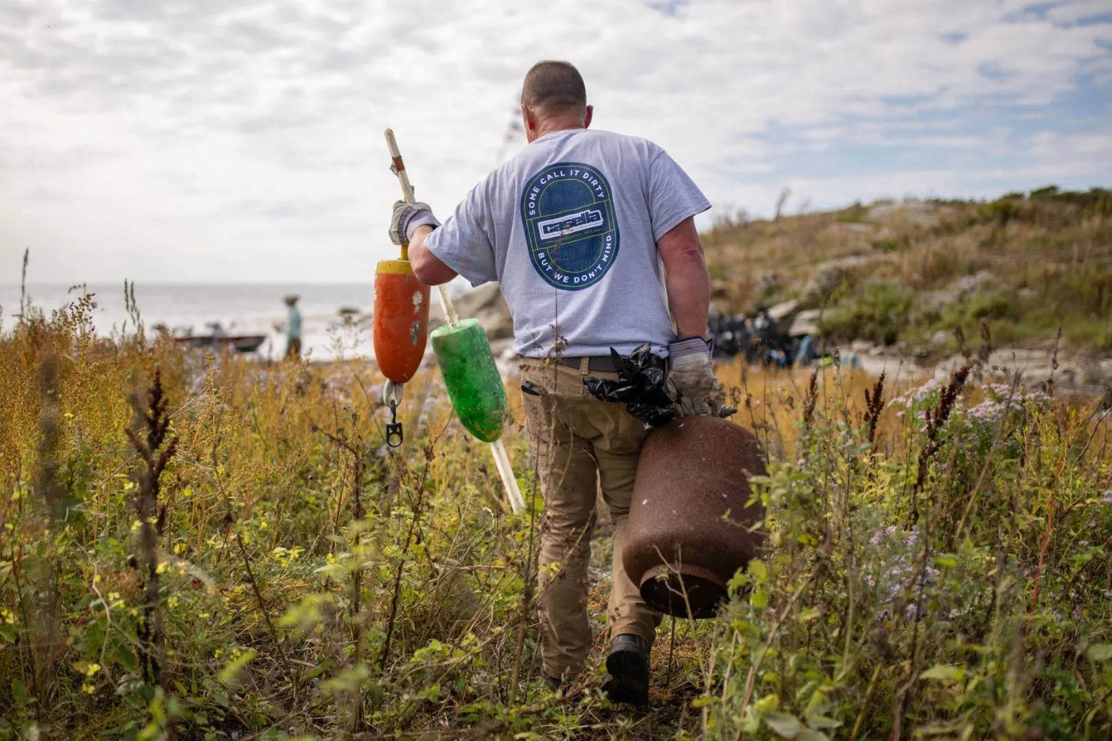 Person wearing a gray T-shirt with a Casella logo on the back walks through tall grass near a rocky shoreline, carrying three colorful buoys and marine debris. The ocean and cloudy sky are visible in the background, with boats anchored offshore.