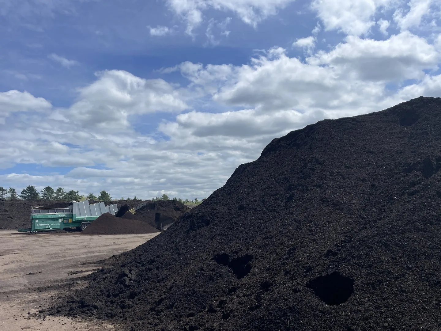 Large piles of dark compost material at an outdoor processing site, with a screening machine in the background under a partly cloudy sky.
