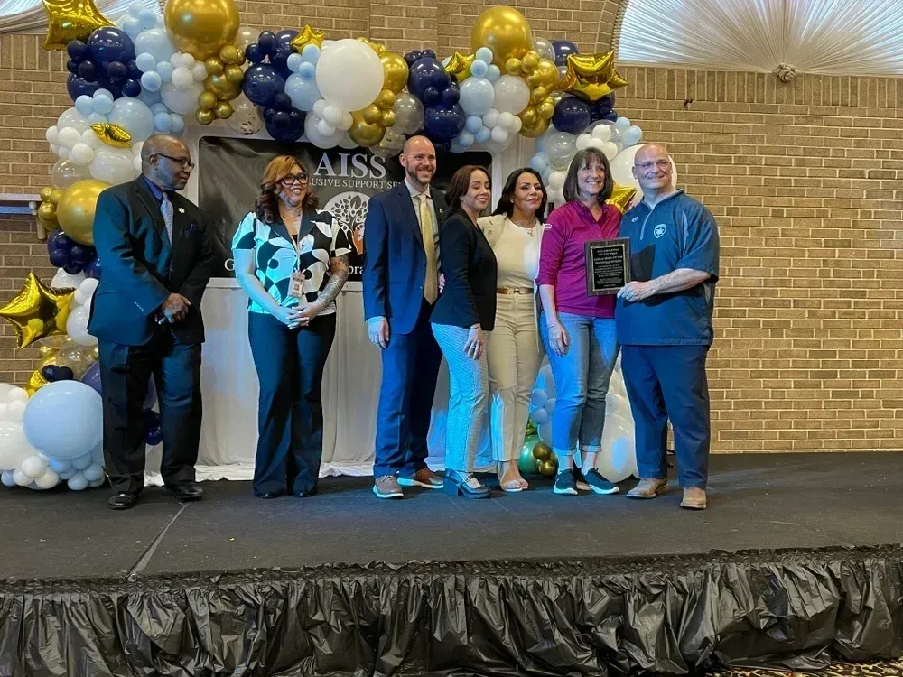 Group of six people standing on a decorated stage with a backdrop of gold, white, and blue balloons arranged in clusters. One person holds a black plaque with silver text, indicating an award presentation. Behind the group is a banner partially visible with the text “AISS” and a tree graphic. The stage has a black skirt, and the background features tan brick walls and white draped fabric overhead.