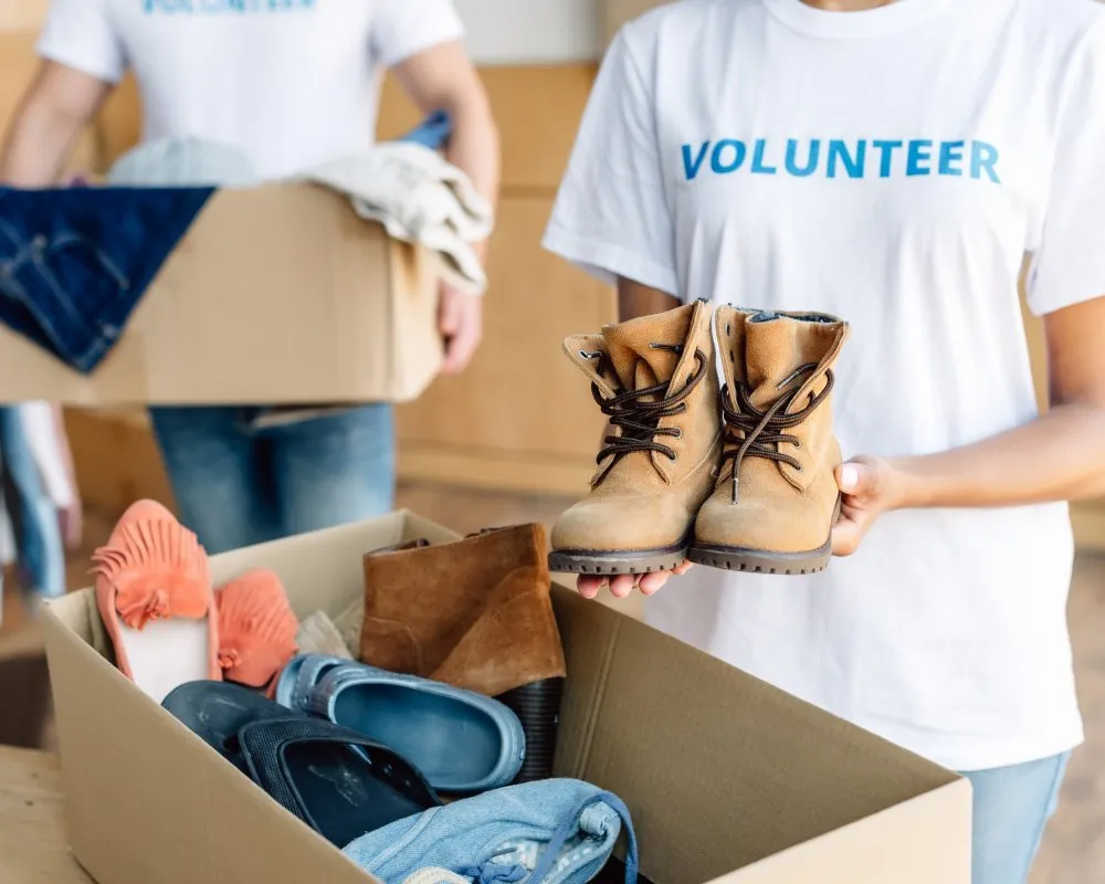 Two volunteers wearing white T-shirts with the word “VOLUNTEER” printed in blue sort donated clothing and footwear. One person holds a pair of tan lace-up boots, while the other carries a cardboard box filled with folded jeans and other garments. A second box in the foreground contains assorted shoes, including sandals, sneakers, and boots, along with folded clothing items. The setting appears to be an indoor donation center with wooden surfaces and racks of clothes in the background. 