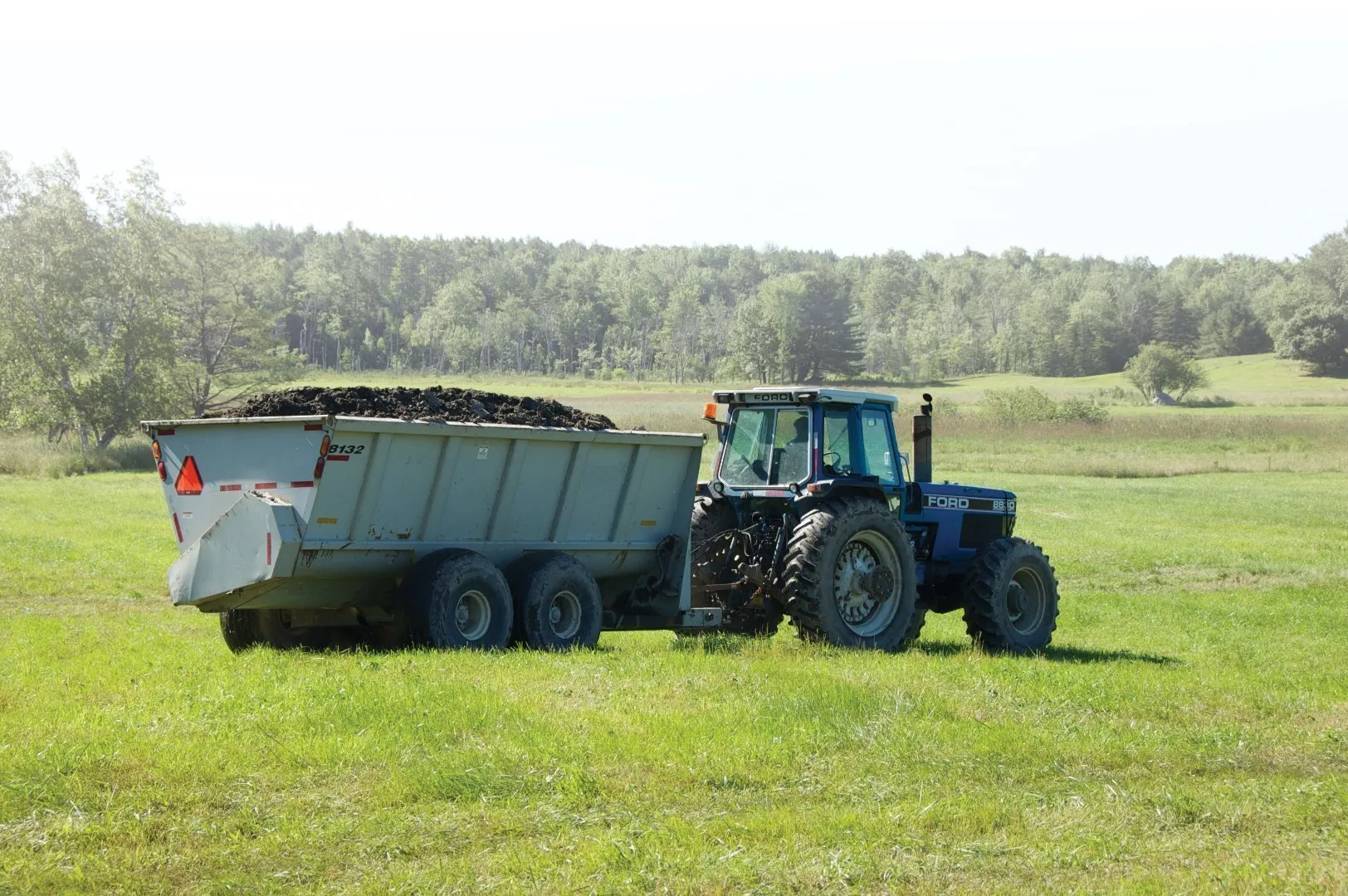 Blue tractor pulling a large trailer loaded with dark compost or soil material across a grassy field, with trees and open farmland in the background under a bright sky.