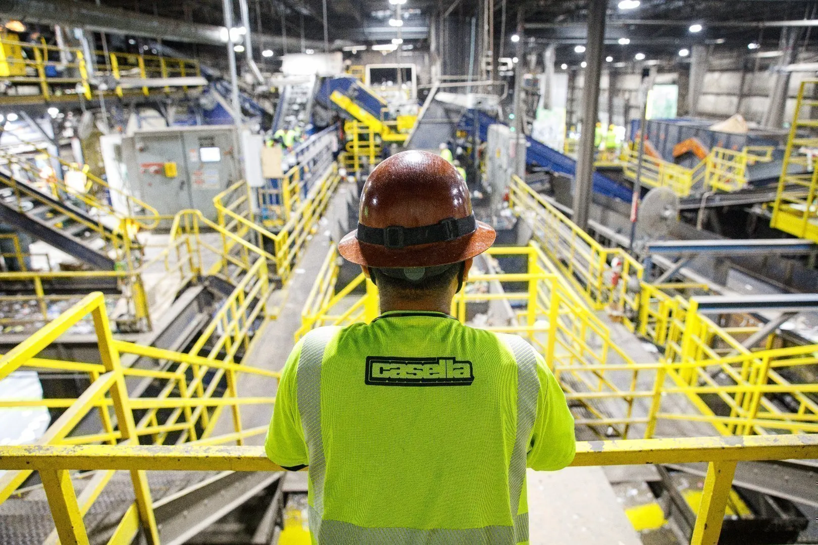 Worker in a high‑visibility Casella shirt and hard hat overseeing operations inside a large recycling facility.