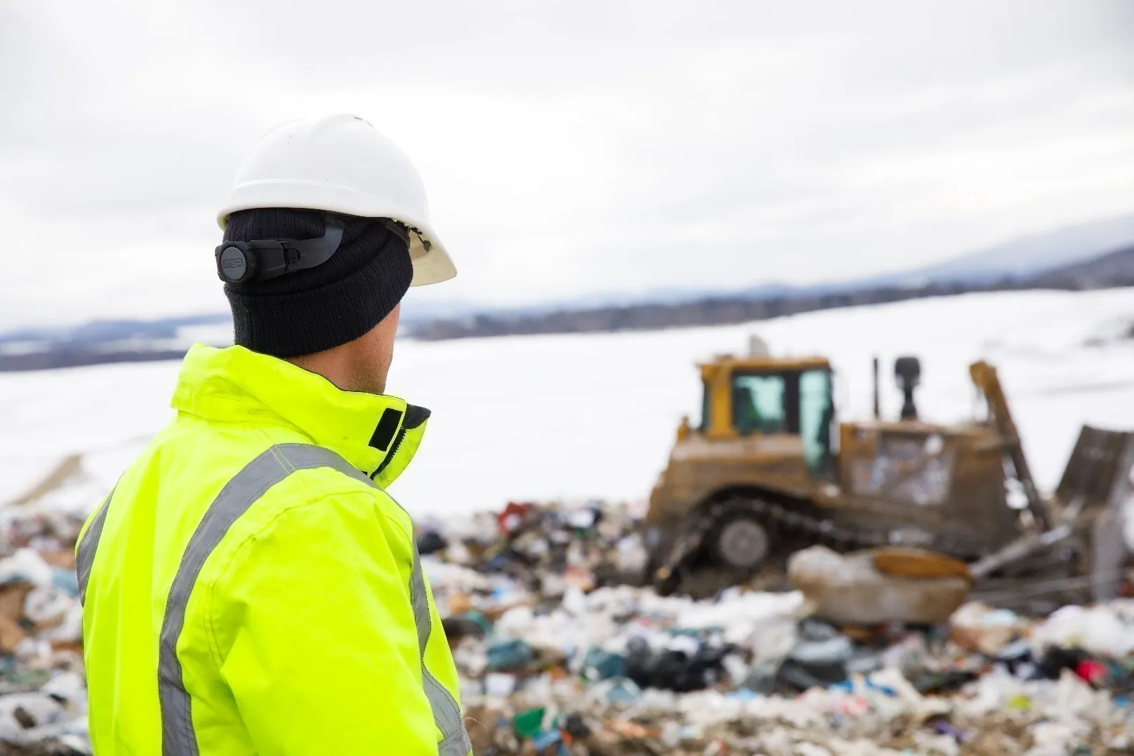 Landfill Environmental Technician wearing a white hard hat and bright yellow high-visibility jacket stands at a landfill site, looking toward a bulldozer moving waste across snowy ground with mountains in the background.