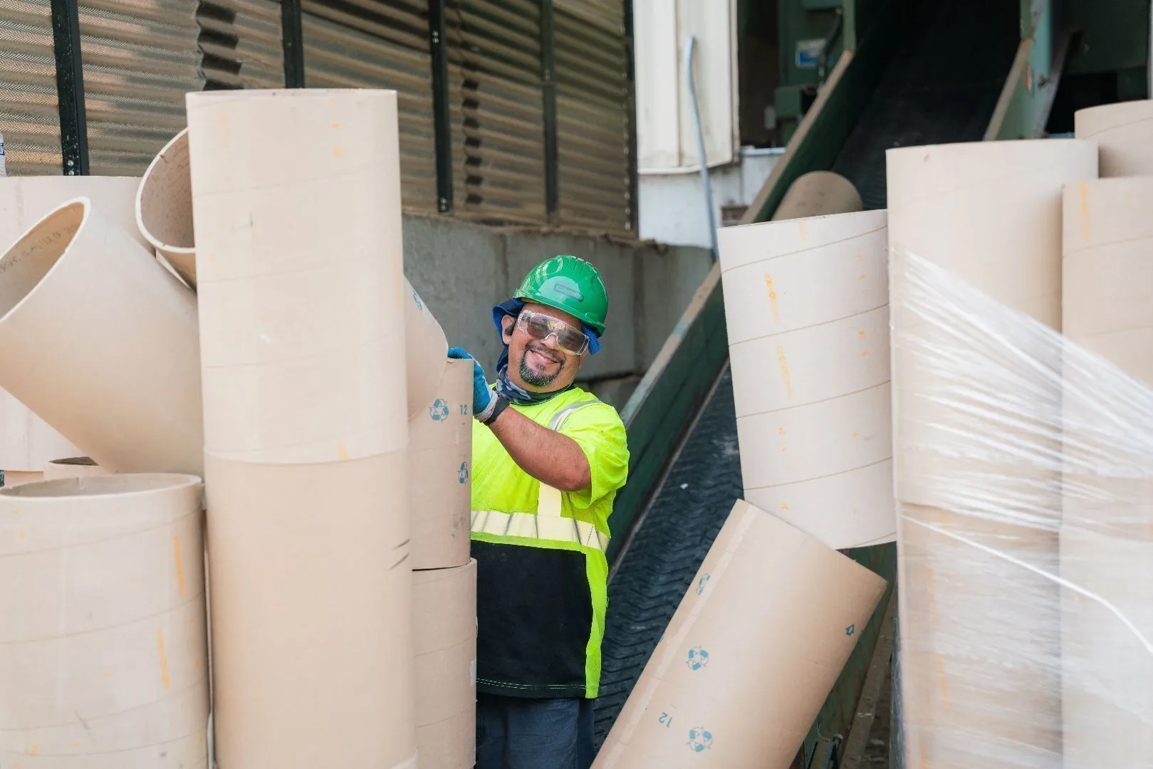 Worker wearing a green hard hat, bright yellow high-visibility safety shirt, and blue gloves handles large cylindrical core tubes near a conveyor belt inside an industrial recycling facility. Several cores are stacked vertically and wrapped in plastic, while others are positioned on the floor.