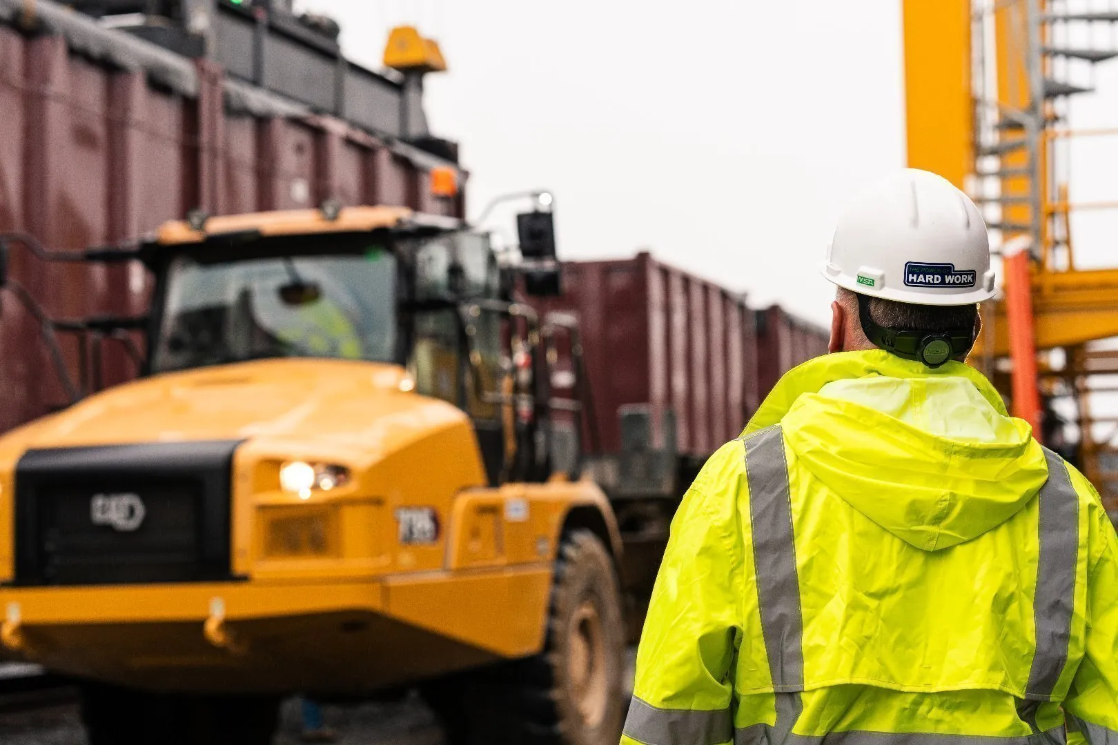 Construction Engineer wearing a white hard hat and a bright yellow high-visibility jacket stands near yellow heavy machinery and maroon rail cars at a Casella landfill site.