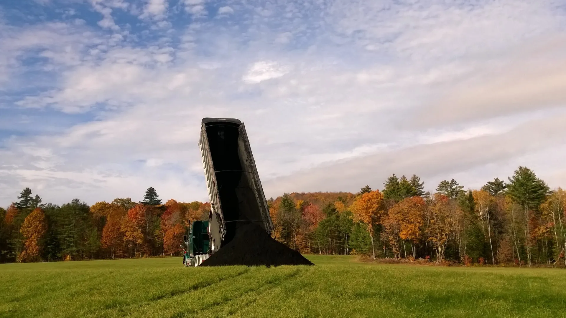Dump truck unloading a large pile of dark material onto a grassy field, with colorful autumn trees and a partly cloudy sky in the background.