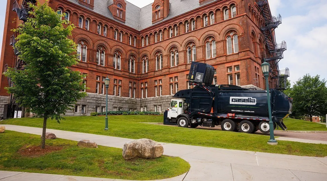 Casella front-load truck lifting a commercial dumpster beside a recycling bin in a tree-lined paved area.