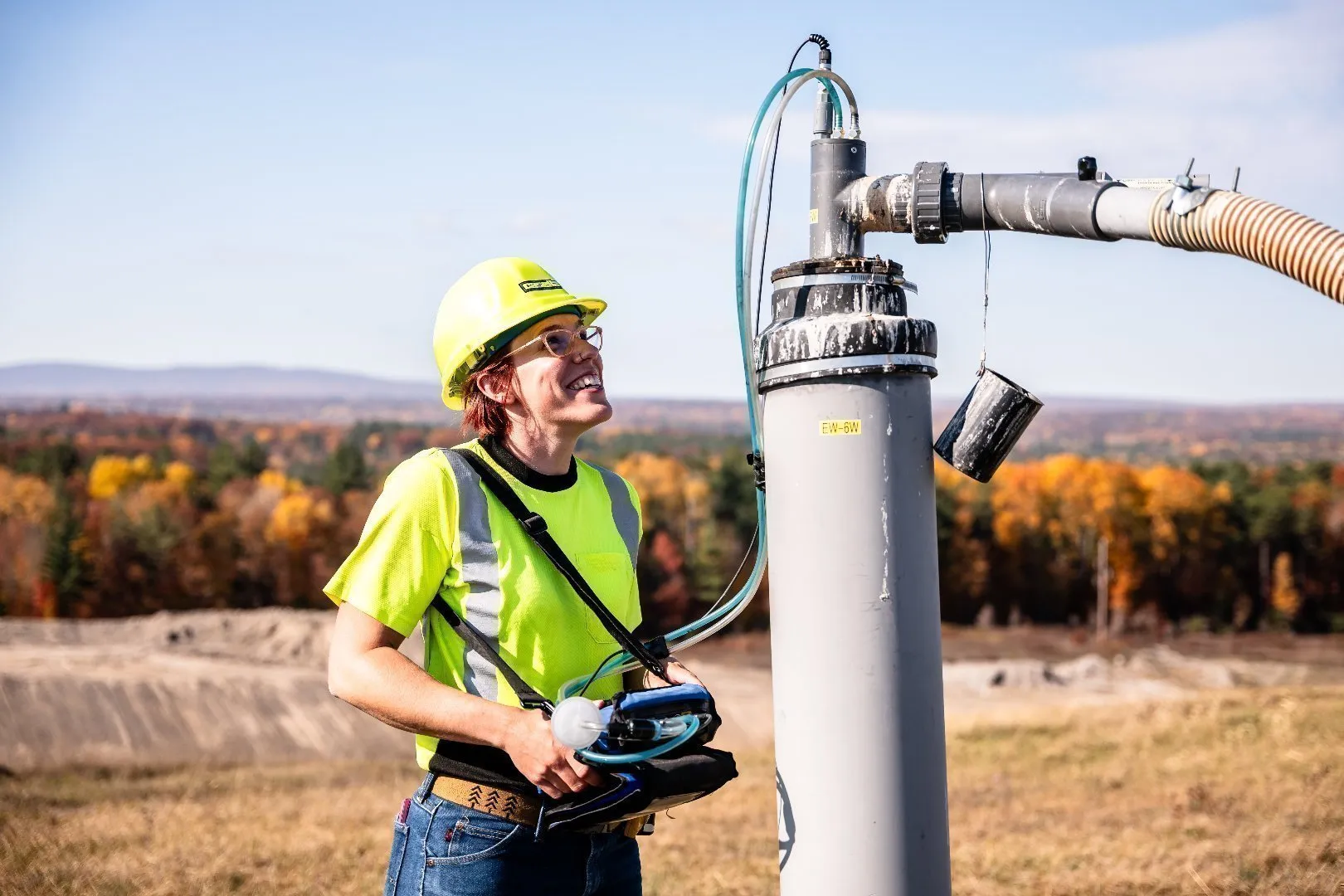 Worker wearing a bright yellow high-visibility safety shirt and hard hat stands outdoors next to a vertical gas well pipe with attached hoses and valves at a landfill site.