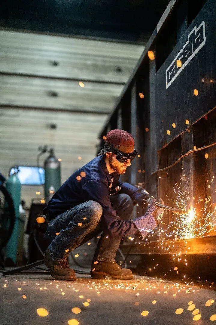 Mechanic wearing protective gloves and a cap uses a welding torch to cut or repair metal on a large Casella dumpster inside a workshop.