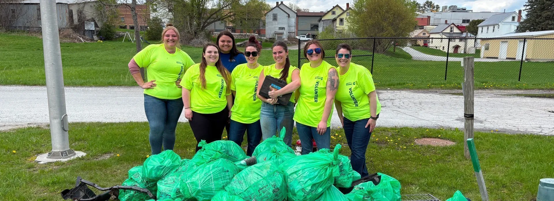 Cleanup volunteers gathered outdoors with a large pile of filled green trash bags, highlighting community recycling and waste‑reduction efforts.