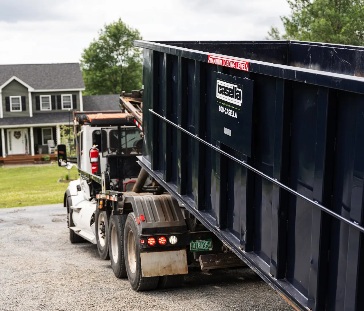 Casella roll-off truck delivering a large dumpster to a residential driveway in front of a two-story home with green lawn and trees.
