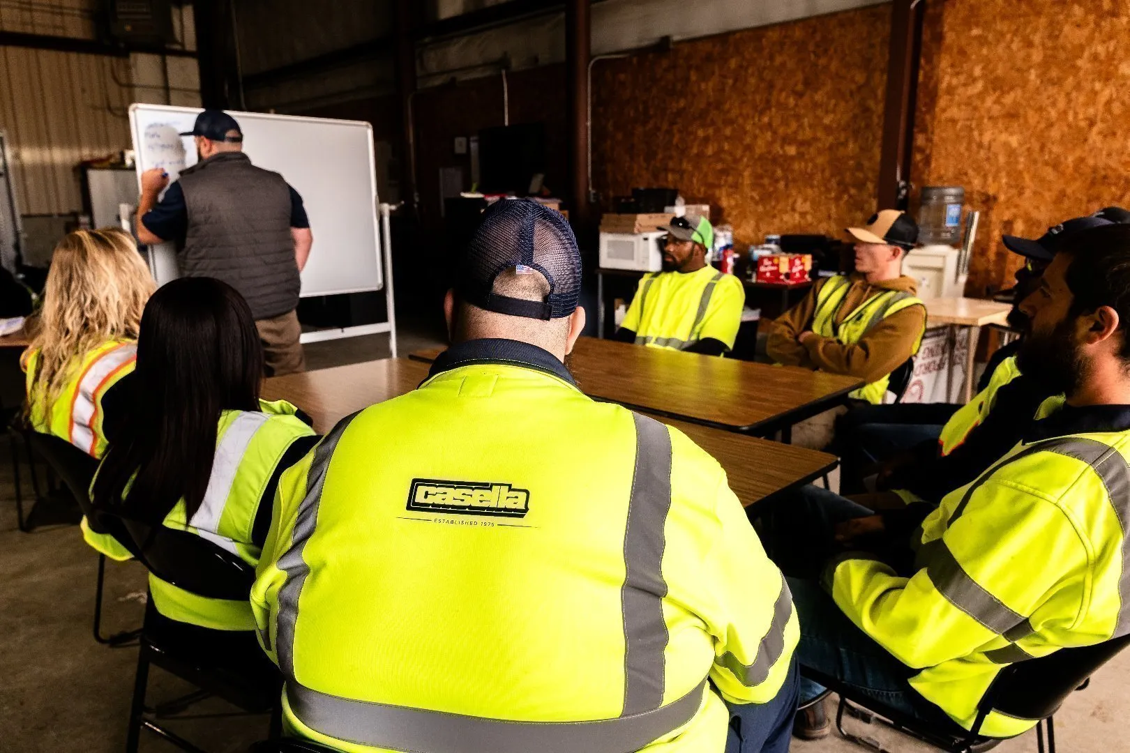 Group of employees in high-visibility safety jackets seated around tables inside a training room while an instructor writes on a whiteboard.