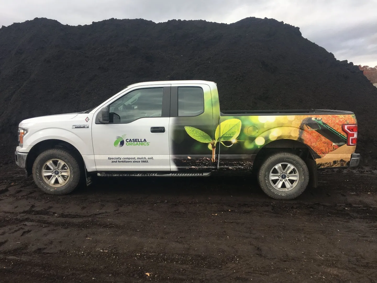 White Casella Organics pickup truck with green plant graphics parked in front of a large pile of dark compost material on a muddy surface.