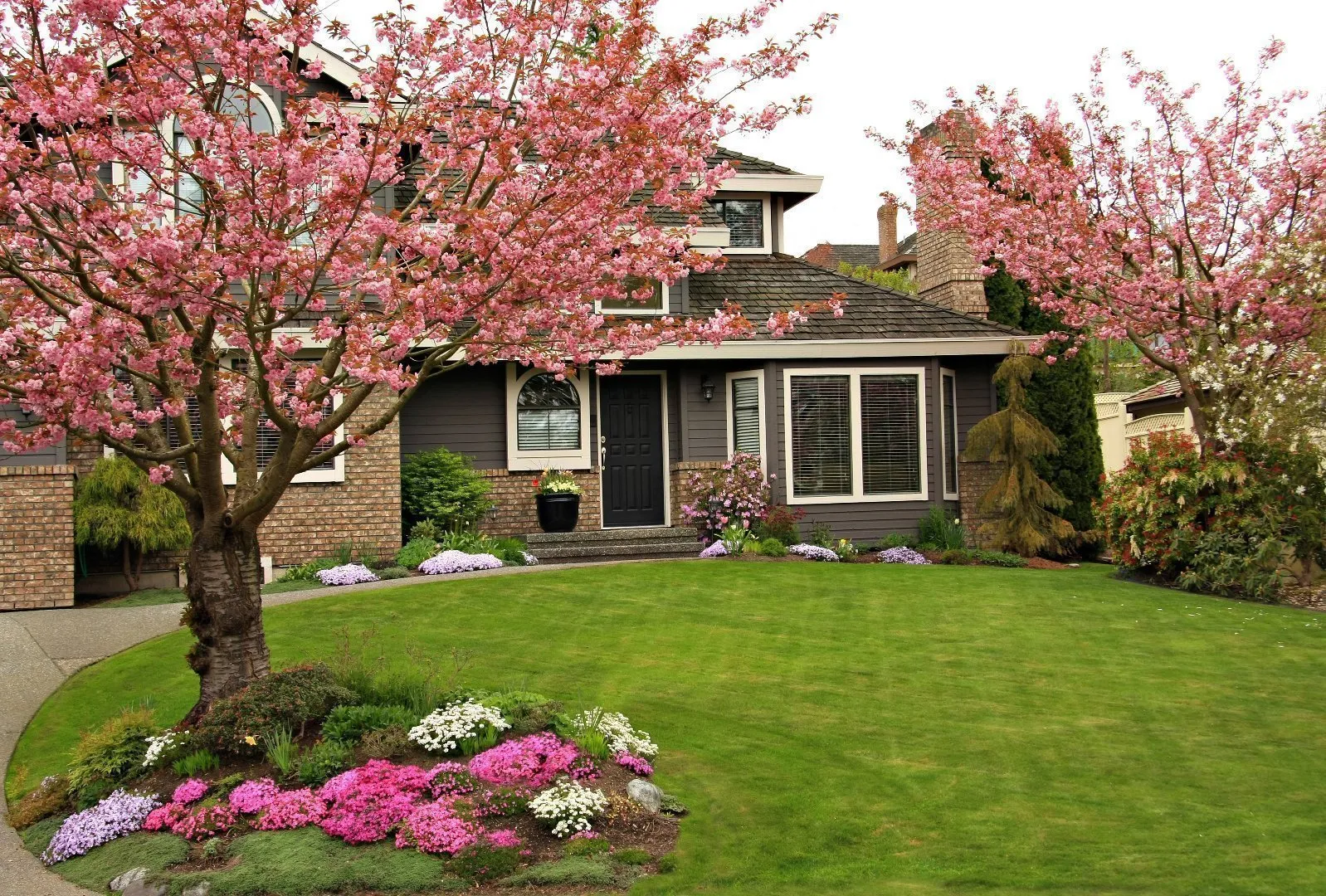 Well-maintained suburban home with a manicured lawn and colorful flower beds, framed by blooming pink ornamental trees in the front yard.