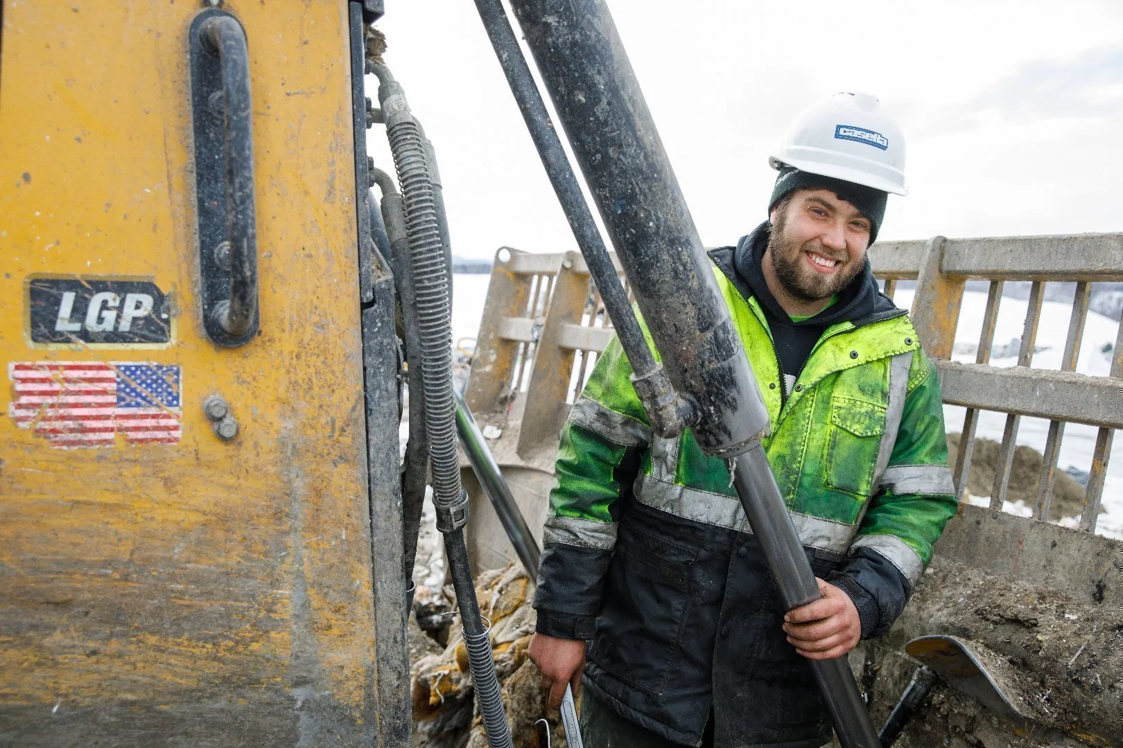 Worker wearing a white hard hat and green high-visibility jacket stands next to heavy equipment with hydraulic arms and hoses.