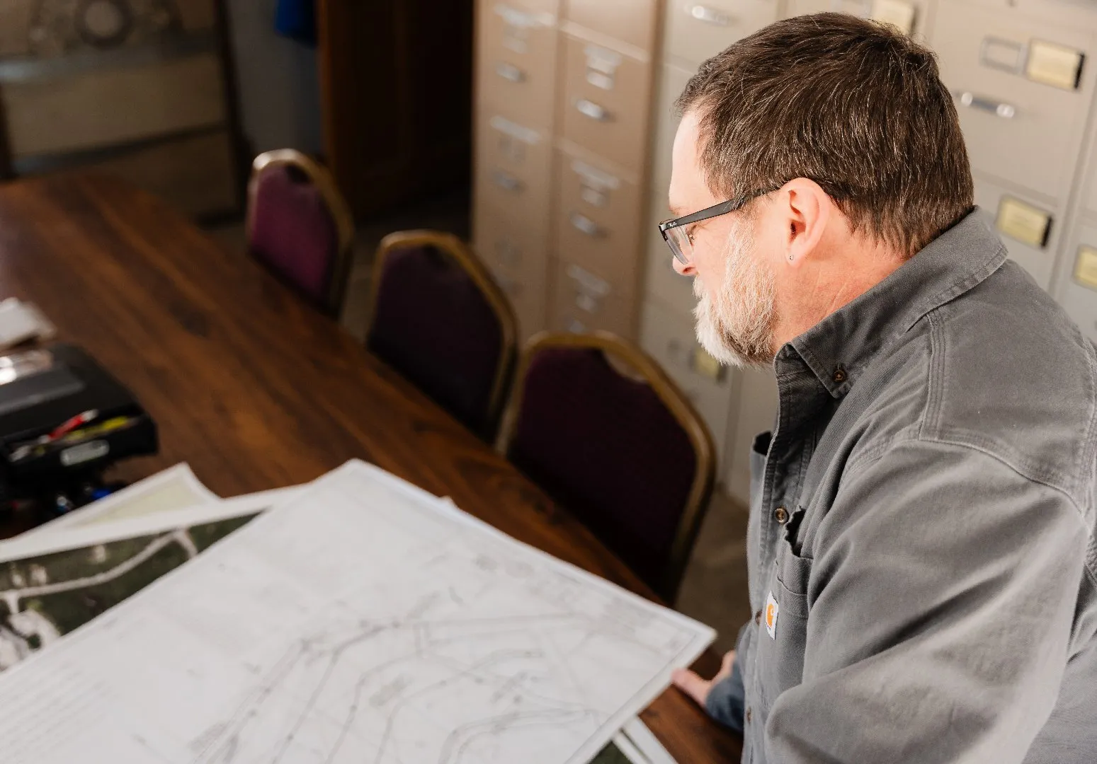 A person reviews large engineering plans on a conference table in an office with filing cabinets in the background.